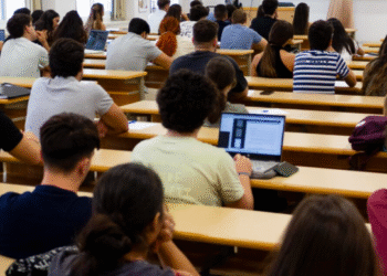 Aula universitaria con estudiantes atendiendo a una clase magistral, algunos tomando apuntes y usando ordenadores portátiles.