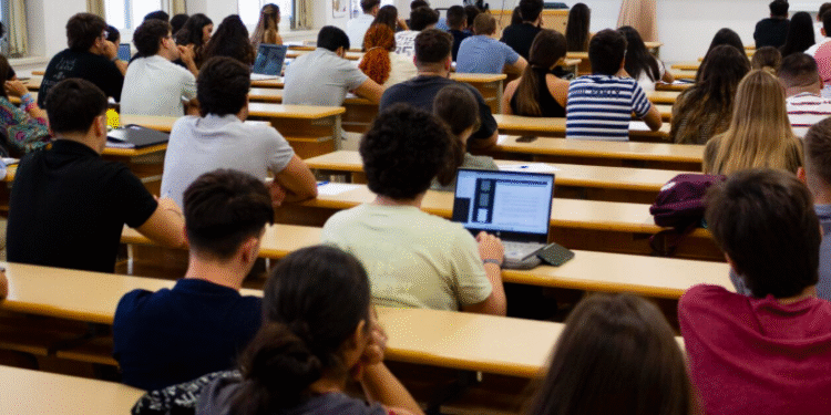 Aula universitaria con estudiantes atendiendo a una clase magistral, algunos tomando apuntes y usando ordenadores portátiles.