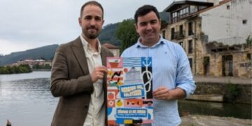 Presentación del cartel del Mercado del Solsticio en Limpias, con dos representantes municipales posando junto a la ría, anunciando la programación de food trucks, talleres y música para el sábado 21 de junio.