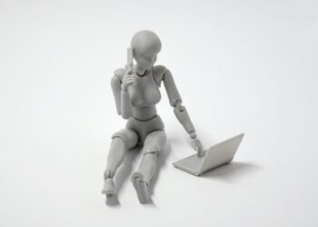 Figura humanoide gris en posición sentada utilizando un ordenador portátil y hablando por teléfono, representando la adaptación tecnológica y organizativa de las empresas a la nueva jornada laboral.